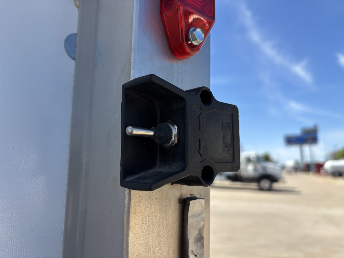 Rear of a 2025 Mack MD7 box truck showing a black power receptacle/connector mount on a silver frame and red taillight, with a blue sky and other vehicles in background.