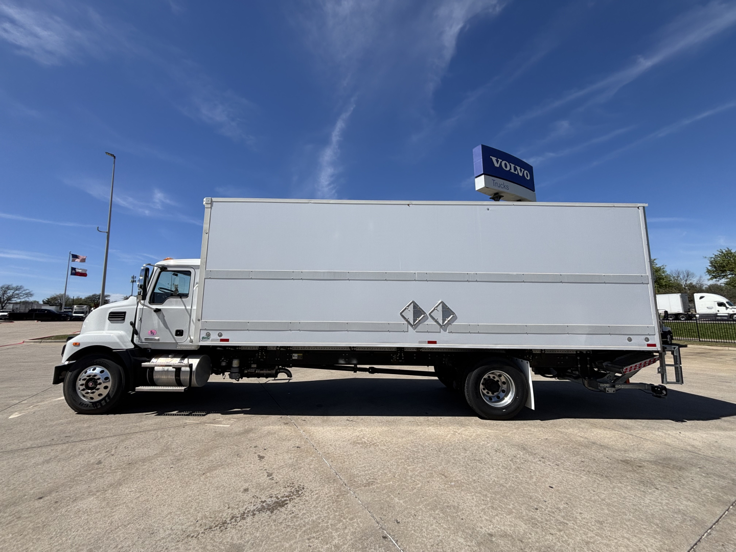 Box truck in side profile showing a large rectangular cargo box on a Mack chassis, white exterior, no text legible on side, blue sky.