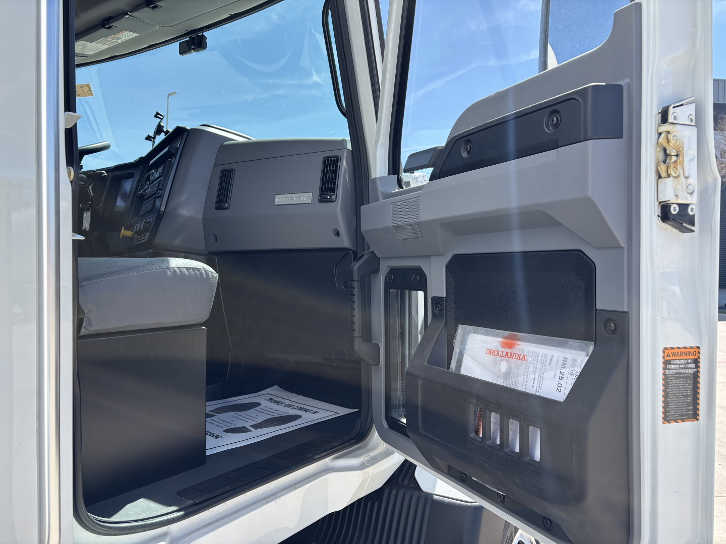 Cab interior of a 2025 Mack MD7 truck showing driver's seat, dashboard, and open door with storage pockets and instructions.