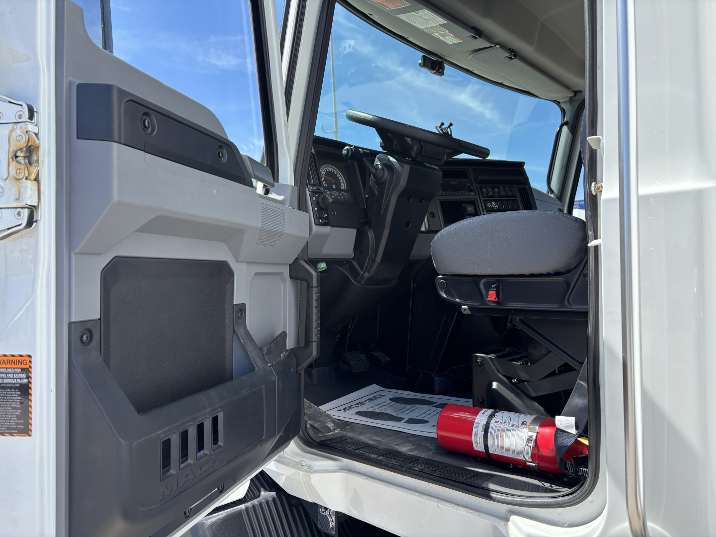 Cab interior of a 2025 Mack MD7 truck showing driver seat, dashboard, steering wheel, and a fire extinguisher in the door sill area; open door