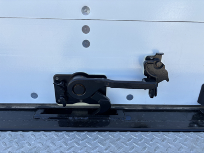 Truck tailgate latch mechanism in view on metal diamond plate, showing latch arm and keeper on a Mack MD7 (new) with VIN tag visible nearby.