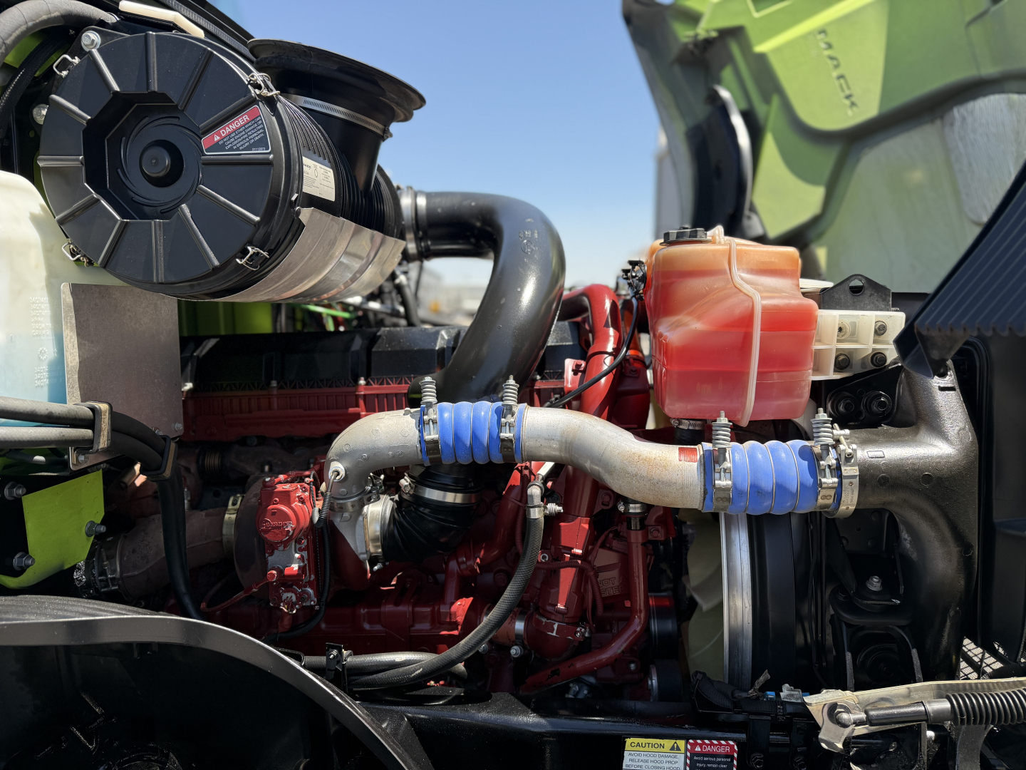 Mack AN64T engine bay showing coolant reservoir, hoses, belts, and turbo manifold. Engine block is red with blue hose fittings and a black radiator hose assembly.