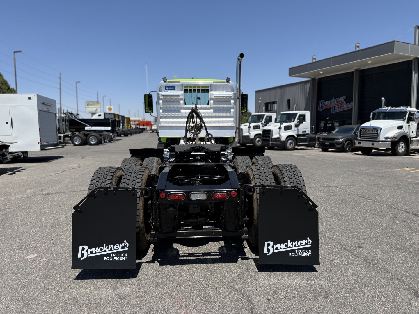 Rear view of a semi truck chassis showing mud flaps with Bruckner's Equipment logos and fifth wheel area, Mack AN64T stock and dealership lot in background.