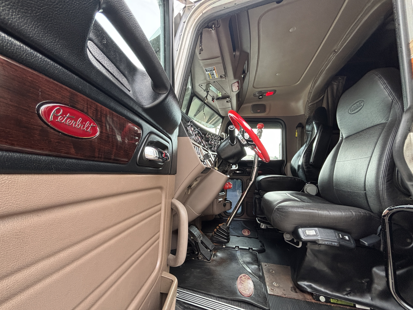 Peterbilt 389 truck interior cab view showing steering column, driver's seat, and dashboard with Peterbilt emblem on door panel.