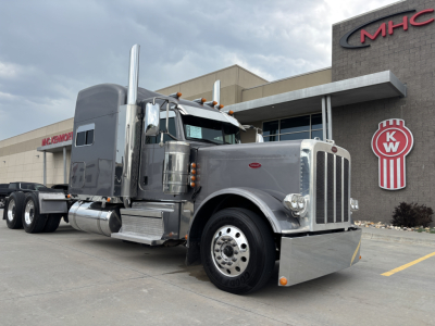 Peterbilt 389 conventional sleeper truck in gray, front three-quarter view, chrome exhaust stacks, large bumper, dual rear axles, parked beside dealership.