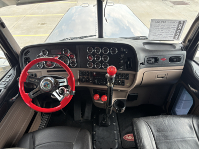 Peterbilt 389 dash and steering wheel interior with red steering wheel and gear shifter; multiple gauges on dashboard; driver seat area visible.
