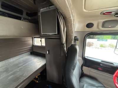 Peterbilt 389 truck interior showing sleeper cab with bunk, cabinet, and mounted monitor/display; driver seat and passenger window visible.