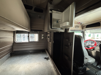 Truck cab interior showing overhead sleeper area with open cabinet and small fridge or storage compartment, and driver seat to the right.