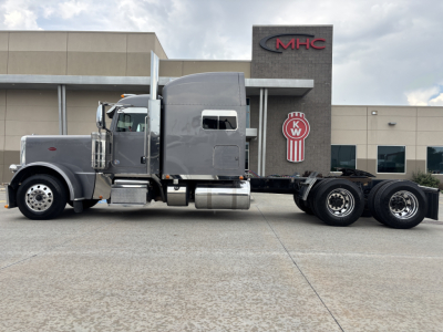 Peterbilt 389 truck in front of a dealership building, gray color, sleeper cab, long chassis, side view with dual rear axles.