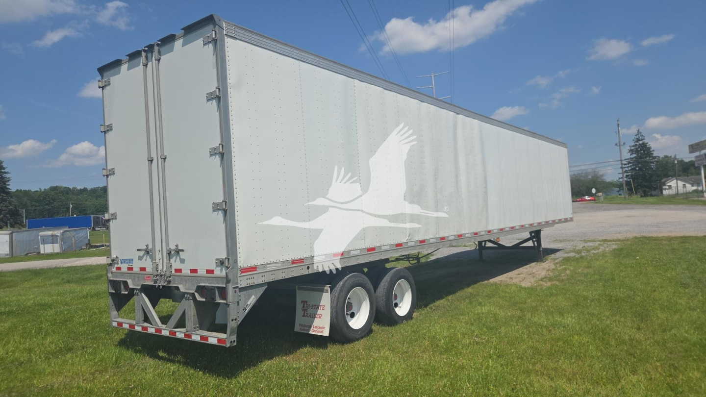 Trailer side view showing white cargo trailer with a stylized bird logo, dual rear doors, tandem axles, and reflective safety stripes.