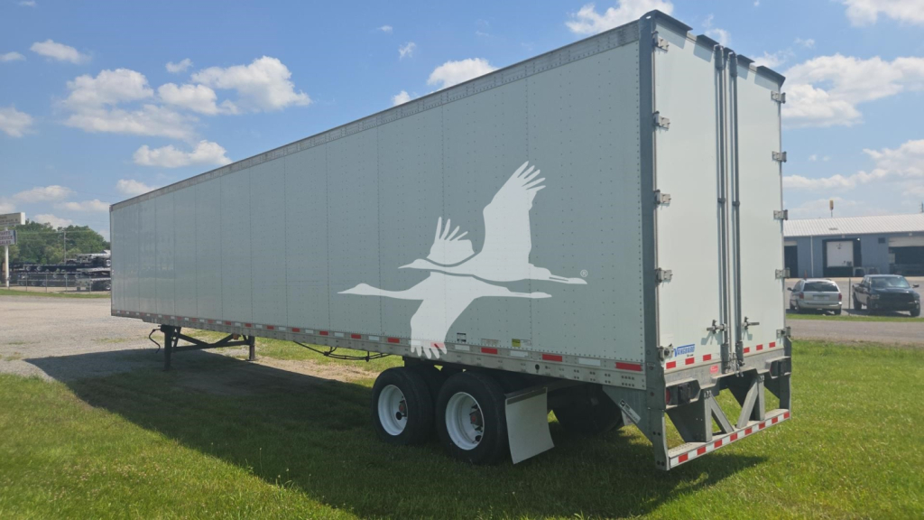 Trailer side view showing a light gray Vanguard trailer with a white bird logo and dual rear doors, flatbed, and tandem axle under bright sky.
