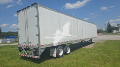 Trailer side view showing white cargo trailer with a stylized bird logo, dual rear doors, tandem axles, and reflective safety stripes.