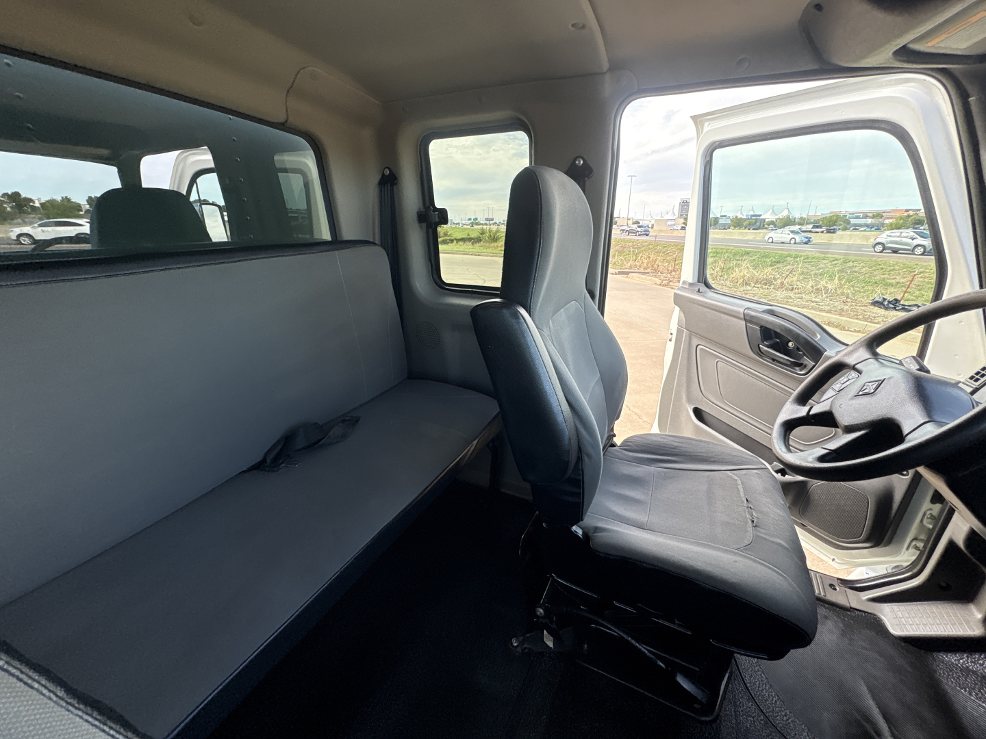 Truck cab interior showing front seat and long bench seat in cargo area. Steering wheel visible, side door open, window view of yard.