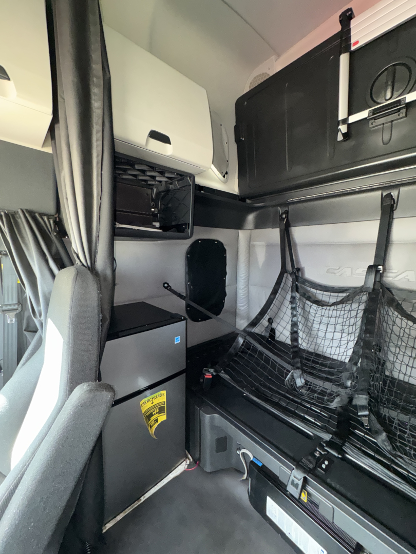 Truck cab interior showing sleeper area with storage cabinets, netting, and compact refrigerator; dashboard area cropped; shelves and black curtain visible.