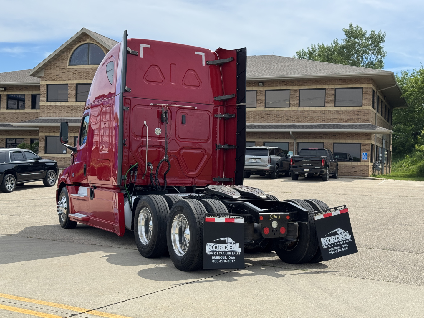 Truck tractor rear three-quarter view showing Freightliner Cascadia 126 sleeper cab in red, with dual rear axles, fifth wheel, and dealership lot in background.
