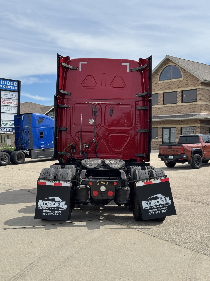 Rear view of a red Freightliner Cascadia tractor with fifth wheel and dual rear axles, mud flaps, and dealership branding on the mud flaps.