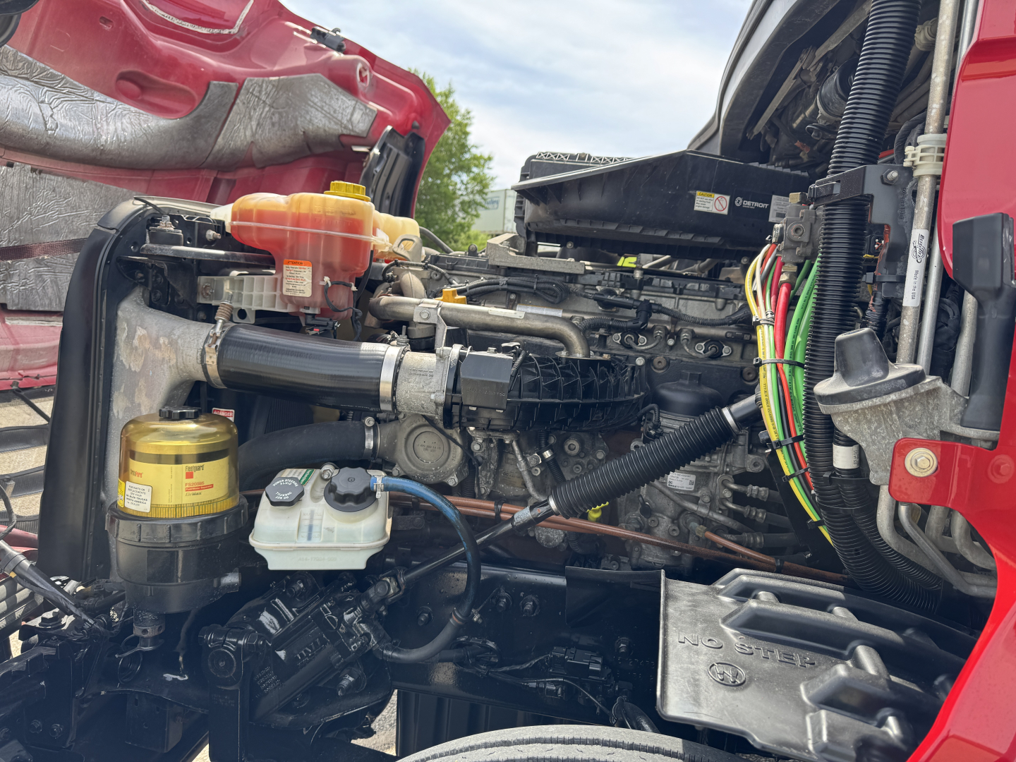 Engine bay of a Freightliner Cascadia showing hydraulic reservoirs, hoses, and wiring harnesses in view.