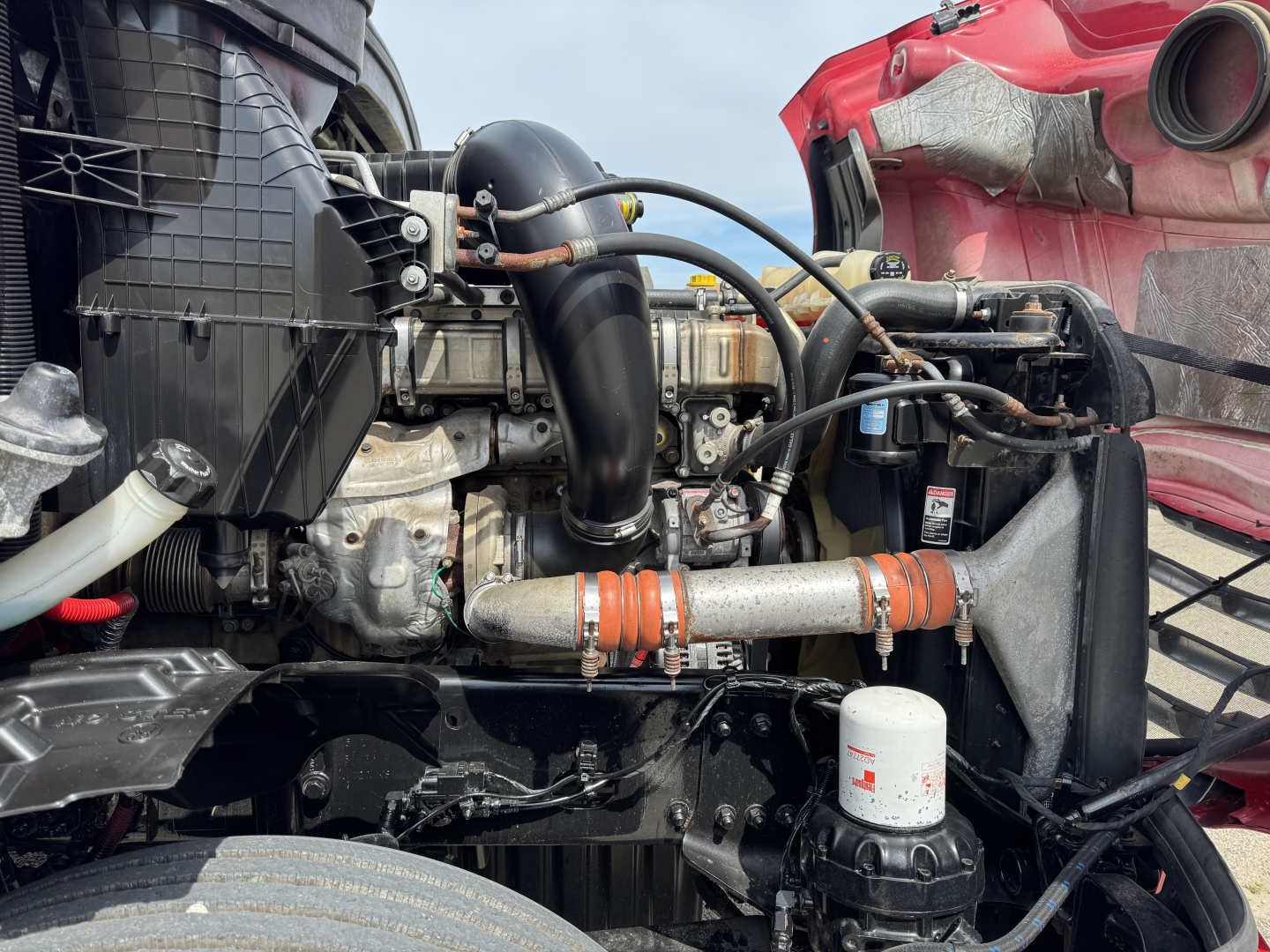 Truck engine bay visible with hoses, radiator, and air intake; multiple black hoses, orange couplings, white filter, and metal piping in front of a Freightliner Cascadia engine bay.