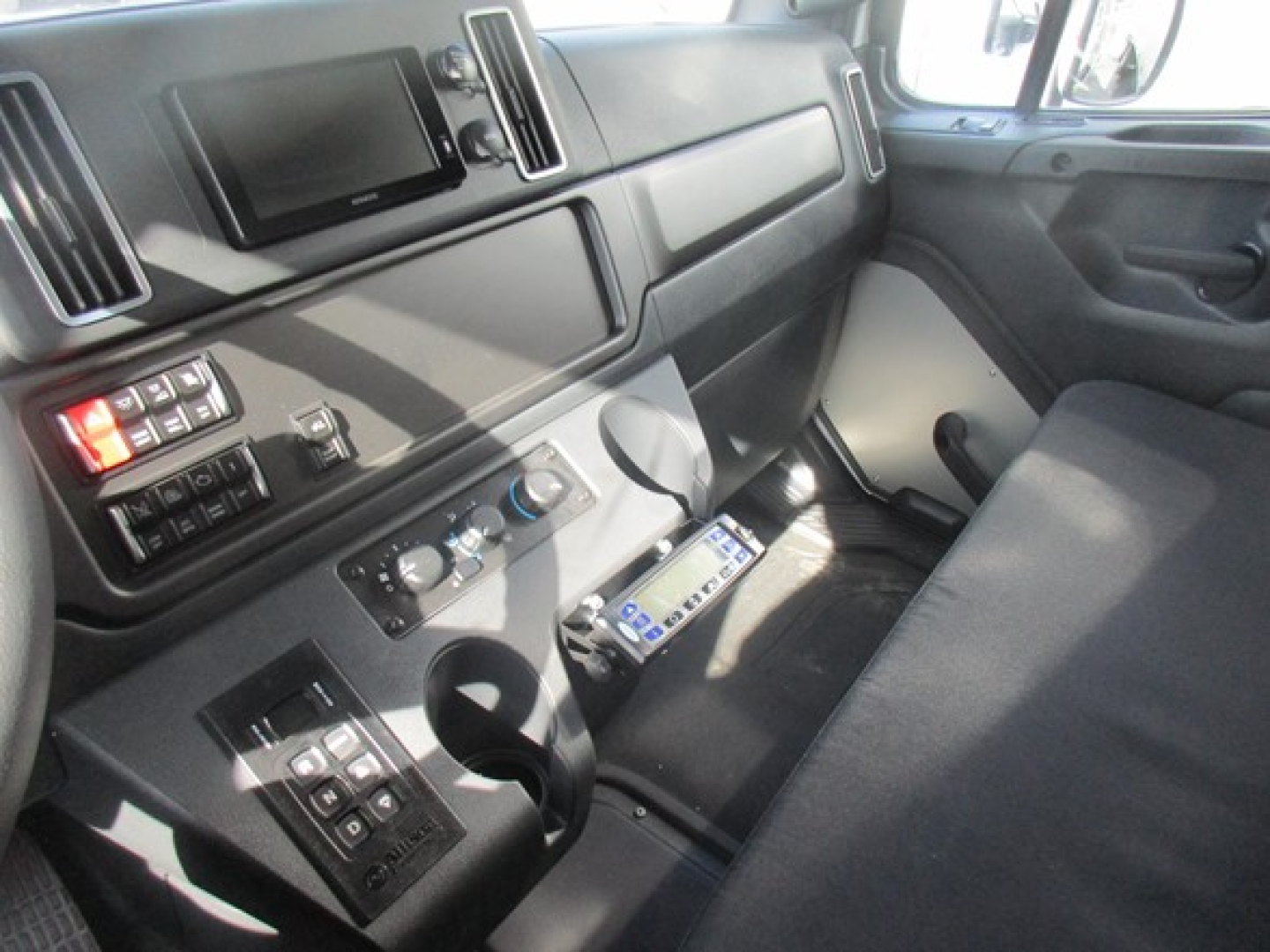 Cab interior of a Freightliner M2 106 Plus showing dashboard controls, steering column, and center console with various switches and a mounted device.