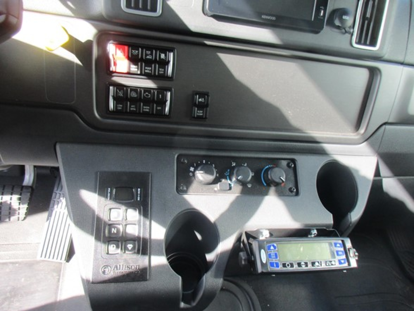 Freightliner M2 106 Plus cabin area showing instrument panel, control switches, cupholders, and a blue device mounted near the center console.