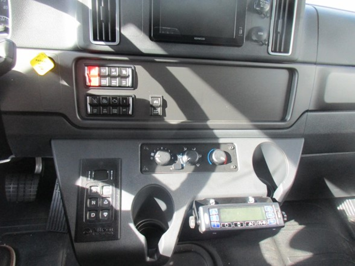 Vehicle interior center console of a 2026 Freightliner M2 106 Plus showing climate controls, cup holders, and a handheld radio unit mounted below the dash.