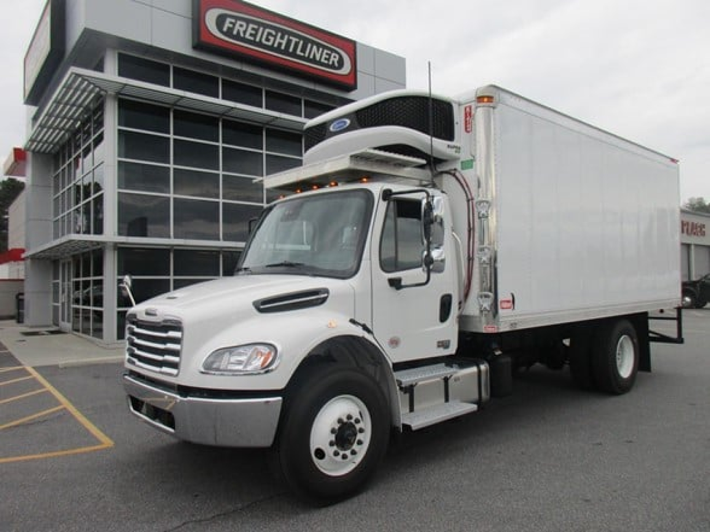 Freightliner M2 106 Plus refrigerated truck side view showing box body and cab in front of dealership.