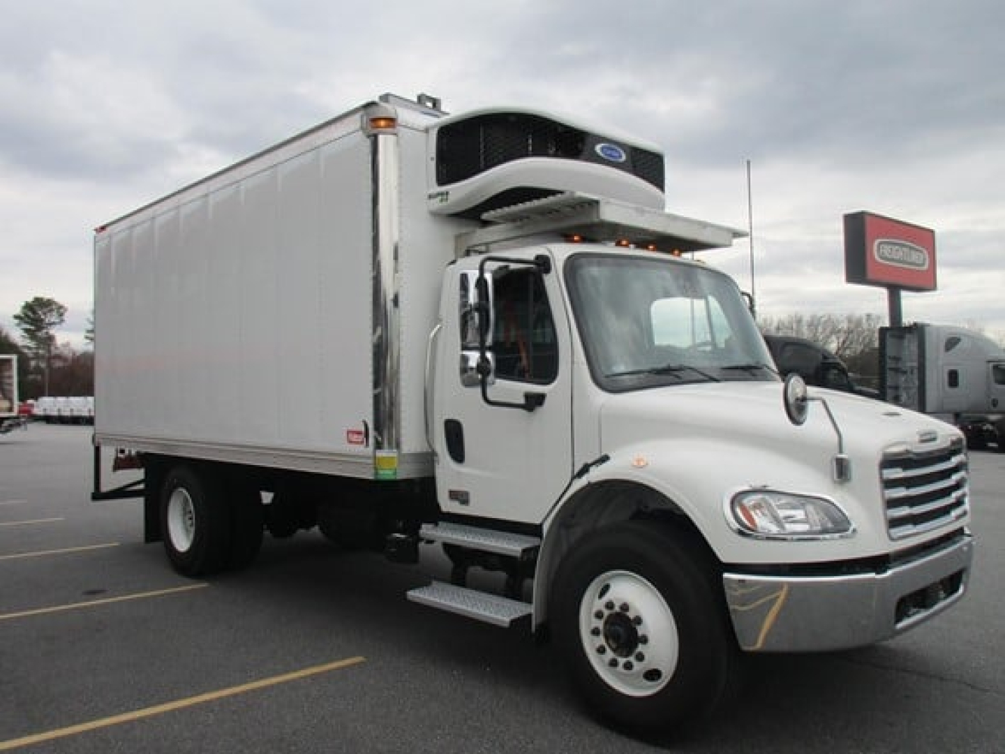 Freightliner M2 106 Plus refrigerated truck with mounted refrigeration unit on top of cargo box, white cab, in-service parking lot.