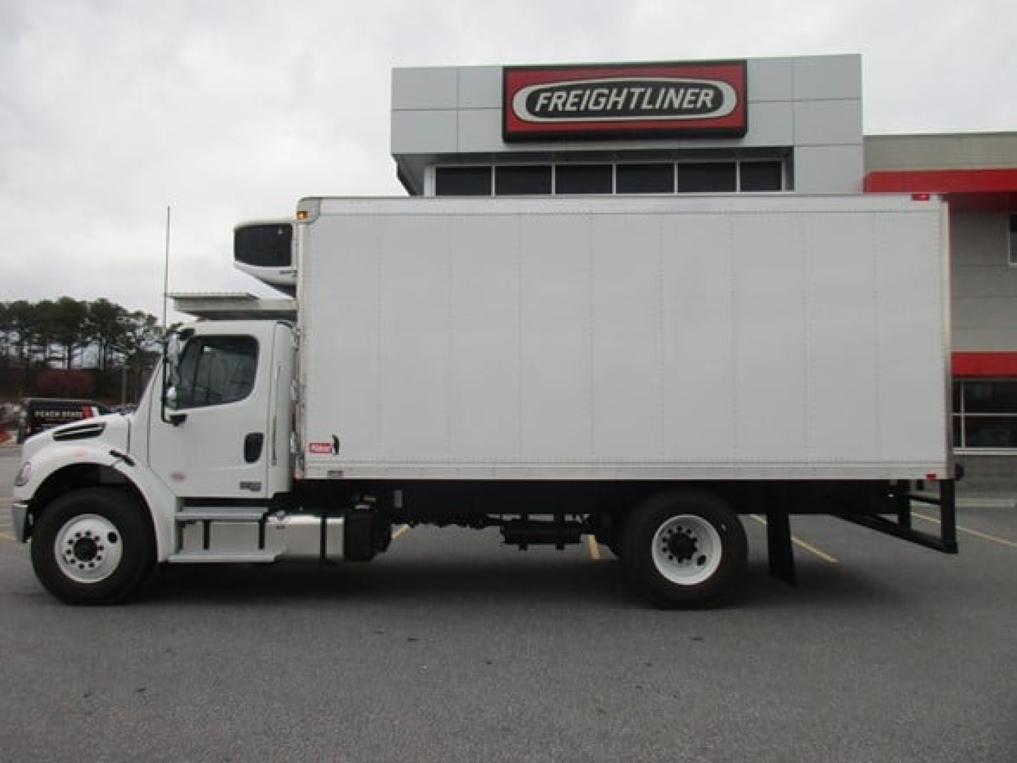 Freightliner M2 106 Plus refrigerated box truck side view parked in front of dealership building.