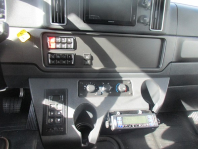 Vehicle interior center console of a 2026 Freightliner M2 106 Plus showing climate controls, cup holders, and a handheld radio unit mounted below the dash.