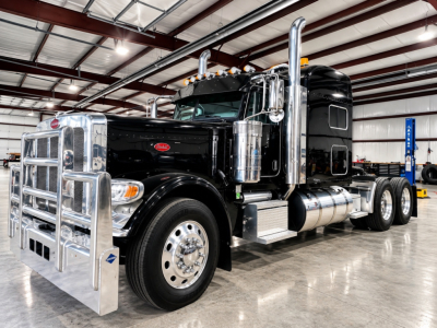 Peterbilt 389 conventional sleeper trucks in a showroom. Front chrome grille with bumper guard, large chrome exhaust stacks, black cab, polished wheels, side chrome fuel tanks.