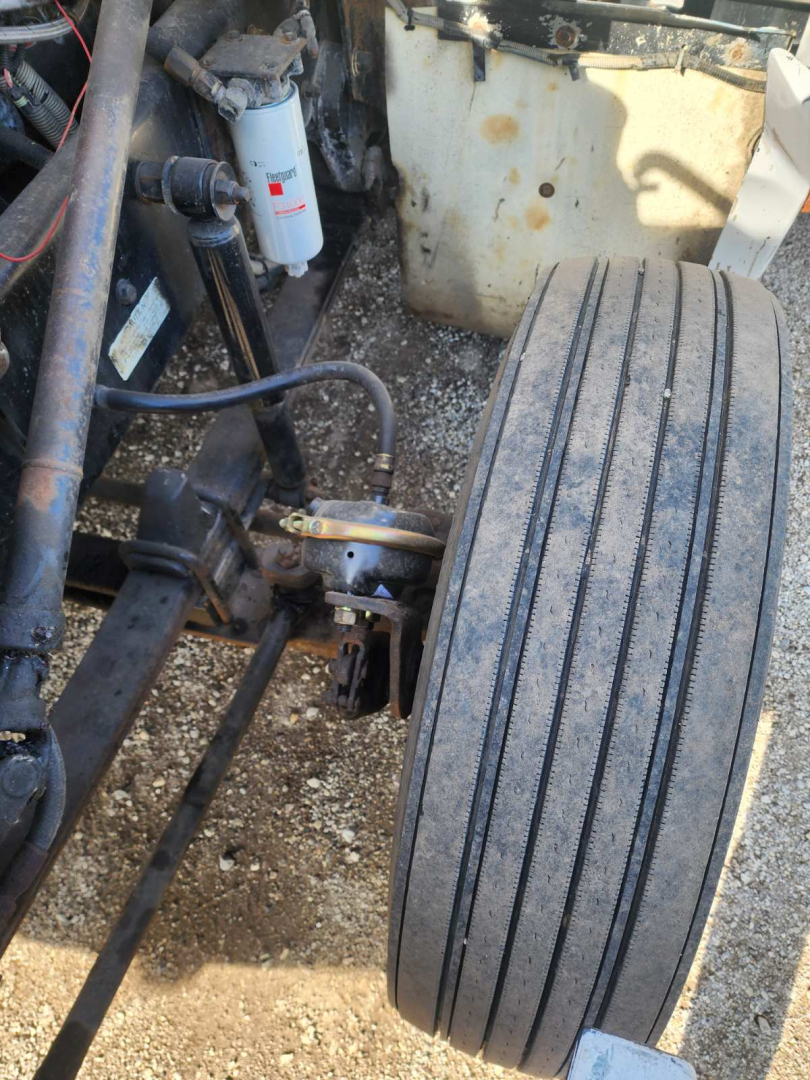 Kenworth T600 front axle area showing dual front tires, suspension components, hoses, and a white filter canister; part of engine bay visible.