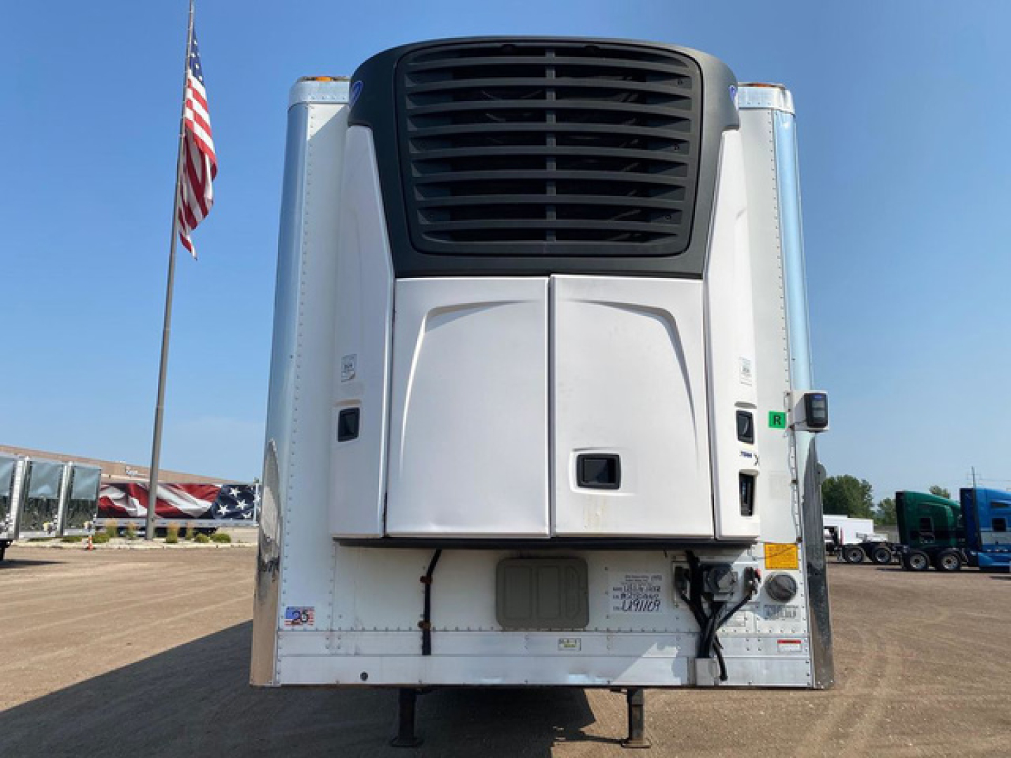 Rear view of a refrigerated trailer with a large black cooling unit at the top and white body, showing undercarriage and connections.