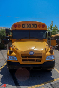 School bus front view showing yellow body, black grille, blue emblem, and 'SCHOOL BUS' sign on the roof; clear blue sky.