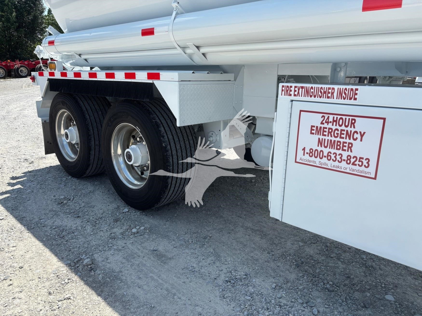Truck trailer side with dual rear wheels and equipment boxes; label reads 24-HOUR EMERGENCY NUMBER 1-800-633-8253; fire extinguisher inside sign visible.