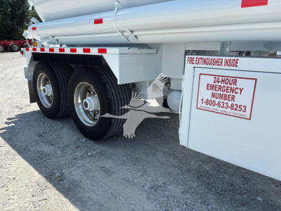 Truck trailer side with dual rear wheels and equipment boxes; label reads 24-HOUR EMERGENCY NUMBER 1-800-633-8253; fire extinguisher inside sign visible.
