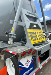 Conquest trailer rear showing yellow license plate style sign "WIDE LOAD" and Conquest branding on gray silos with blue and red logo, part of pneumatic dry bulk trailer setup.