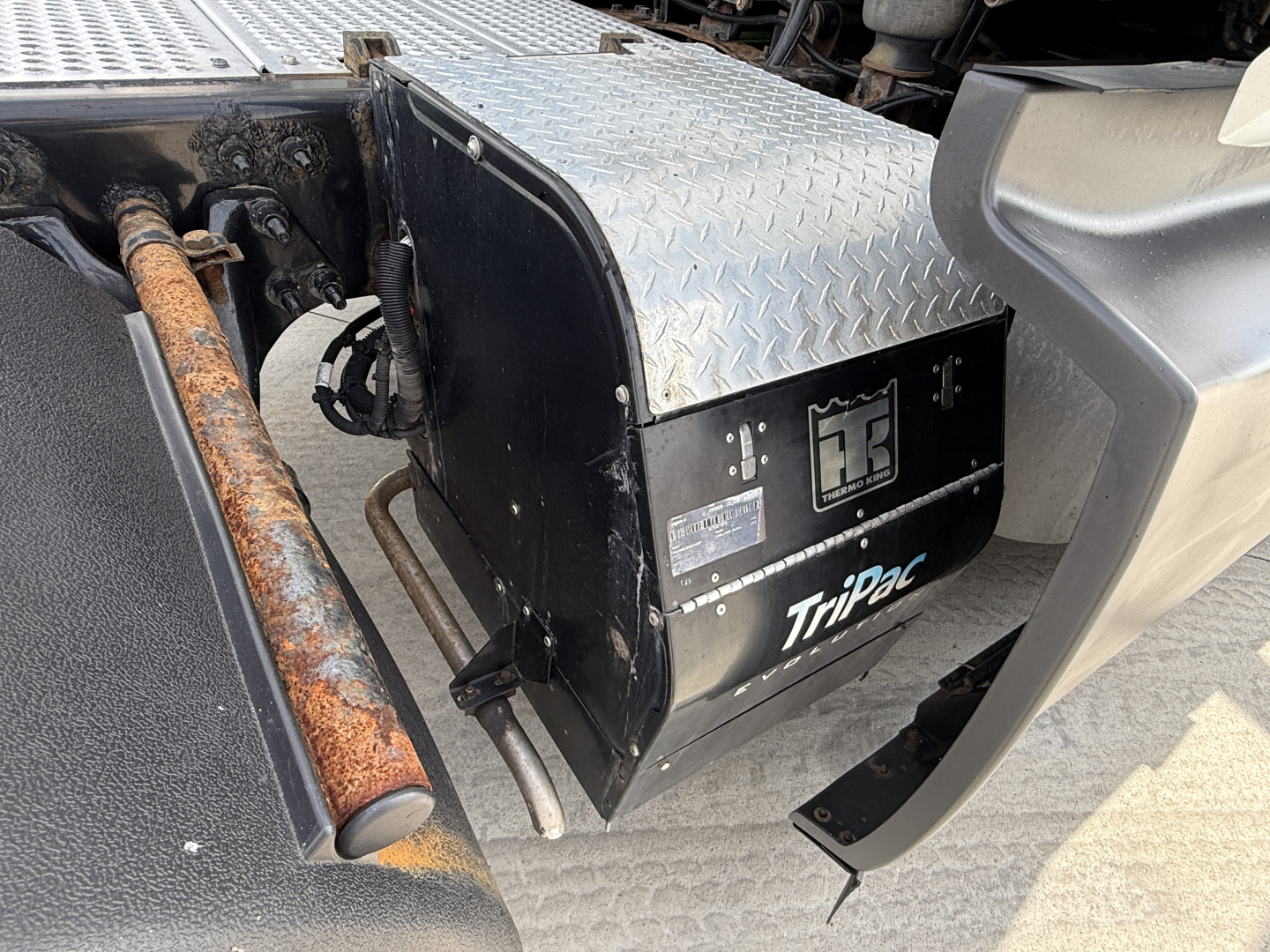Truck saddle area showing a black TriPac auxiliary power unit with Diamond Plate toolbox and rusted exhaust pipe; side steps and cab area visible.