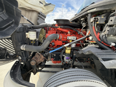 Engine bay showing a red Cummins ISX engine with surrounding hoses, wiring, and a black radiator on the left. Visible components include a white reservoir with text, a black air filter housing, and a small yellow filter. The right side show