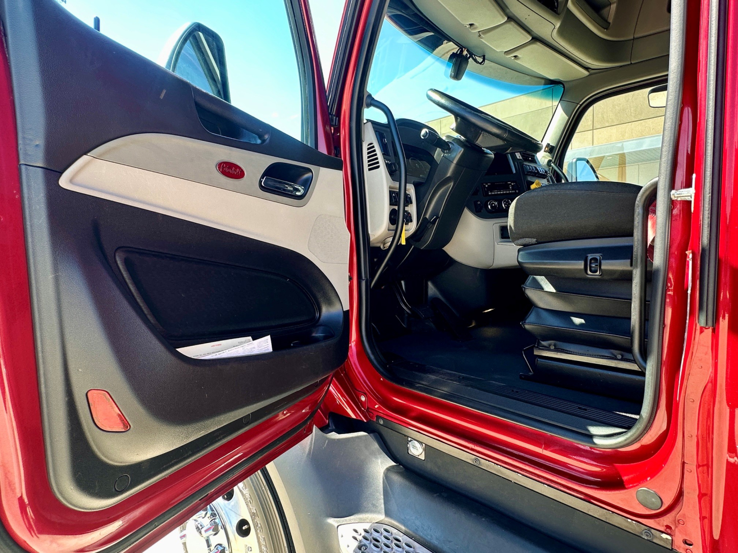 Truck cab interior view of a red 2023 Peterbilt 579 through open driver door, with steering wheel, dashboard, and seating visible.