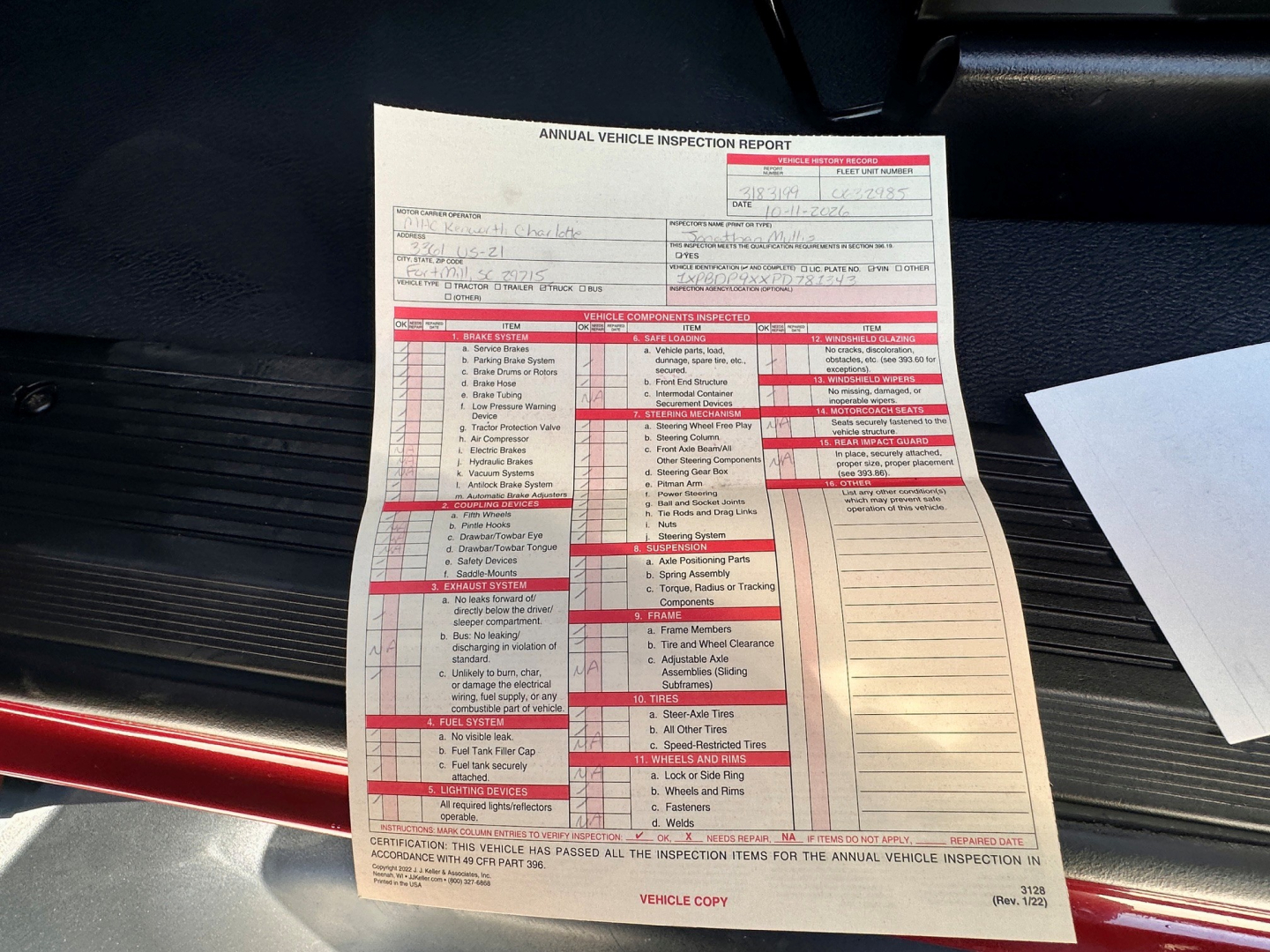 Truck interior showing an Annual Vehicle Inspection Report with red sections and text, placed on the floor mat near the driver side; a white paper with inspection details is visible.