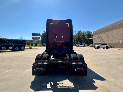 Rear view of a 2023 Peterbilt 579 conventional sleeper truck chassis in a parking lot showing dual rear axles, mud flaps with MHC logo, and red cab in the background.