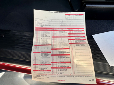 Truck interior showing an Annual Vehicle Inspection Report with red sections and text, placed on the floor mat near the driver side; a white paper with inspection details is visible.