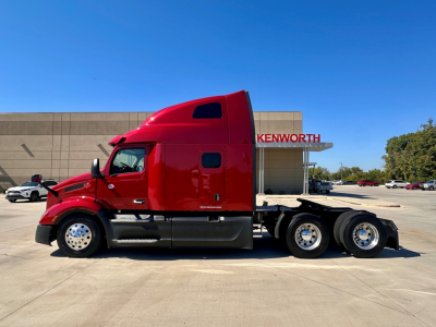Peterbilt 579 conventional sleeper tractor in red, side view with three axles at rear, fuel tanks, and Kenworth dealership in background.