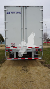 Rear view of a Lange trailer with double doors, River Valley trailer sticker, and reflective red/white safety tape along bumper; two vertical hinges and small decals near lower left.