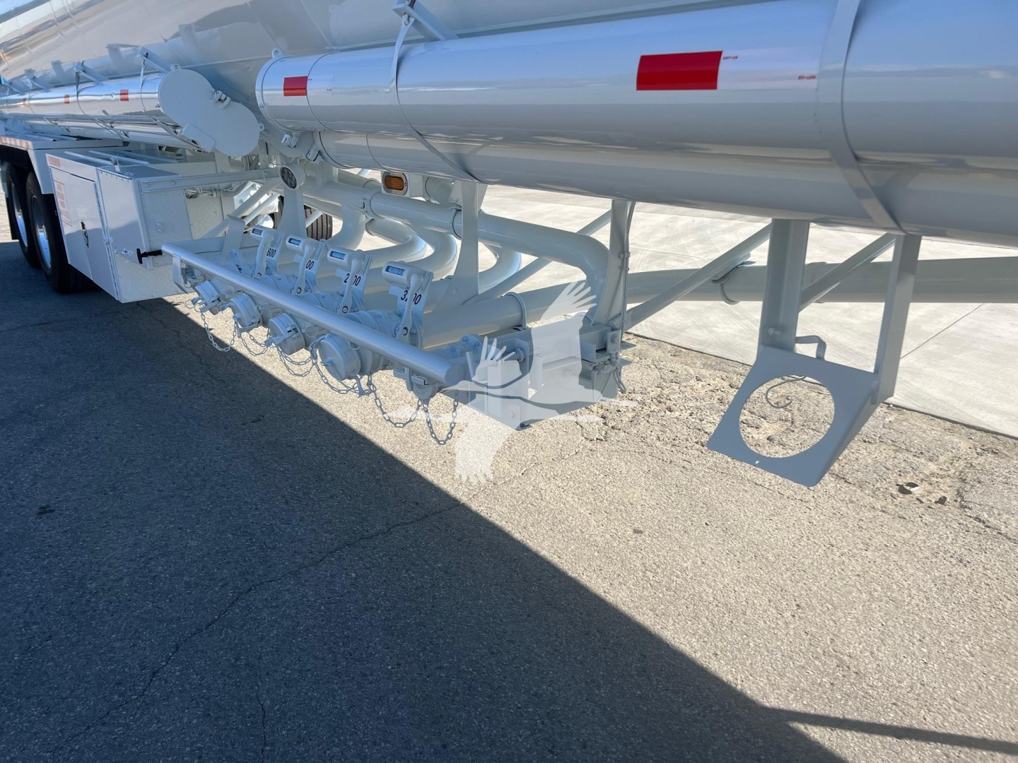Tank trailer undercarriage showing multiple white pipes and valves along side with trailer frame and wheel area visible.