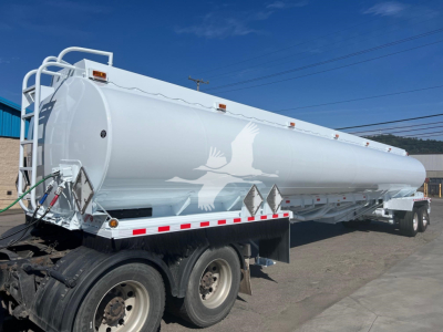 Tank trailer showing white Heil Petro fuel tanker with curved top and safety decals, side-mounted ladders, and reflective striping along lower edge.