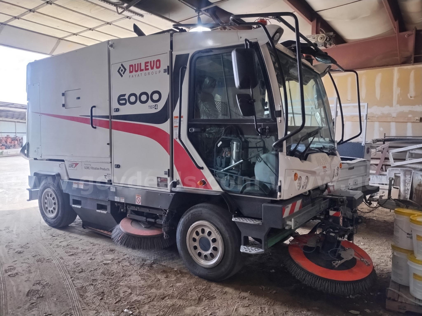 Elgin 6000 sweeper truck in workshop showing front-right view, DULEVO branding, 6000 logo, red and black stripe along cab, spinning cylindrical brushes visible under carriage