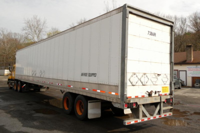 Rear-right side of a silver Vanguard VXP trailer with 3-axle wheels, rear doors, and safety decals; visible text includes ANACEL Co... and diamond placards; rear bumper and reflectors.