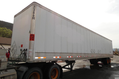 Trailer side view showing a large rectangular white Vanguard VXP trailer with rear wheels, delivery hitch, and orange reflective markers.