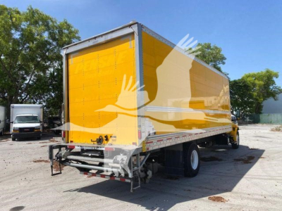 Box truck rear-side view showing bright yellow cargo box with gray underrcarriage and rear bumper; International MV 2022 likely model; visible white cab and open lot background.